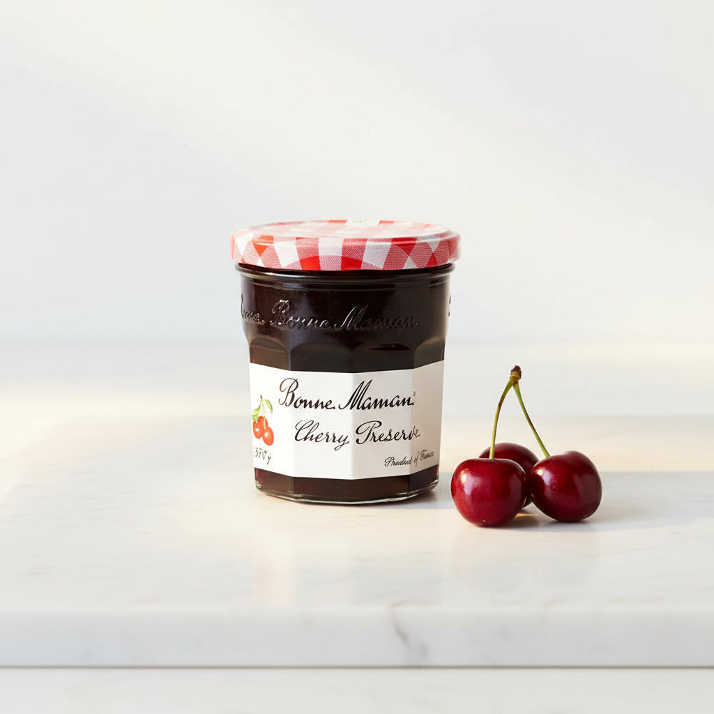 A glass jar of Bonne Maman Cherry Reserves jam with a red and white checkered lid and a white label, set against a plain off-white background.