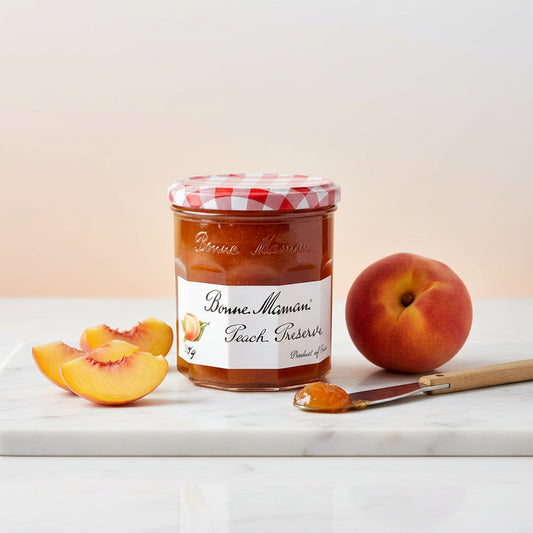 A glass jar of Bonne Maman Peach Reserve jam with a red and white checkered lid and a white label, set against a plain off-white background.